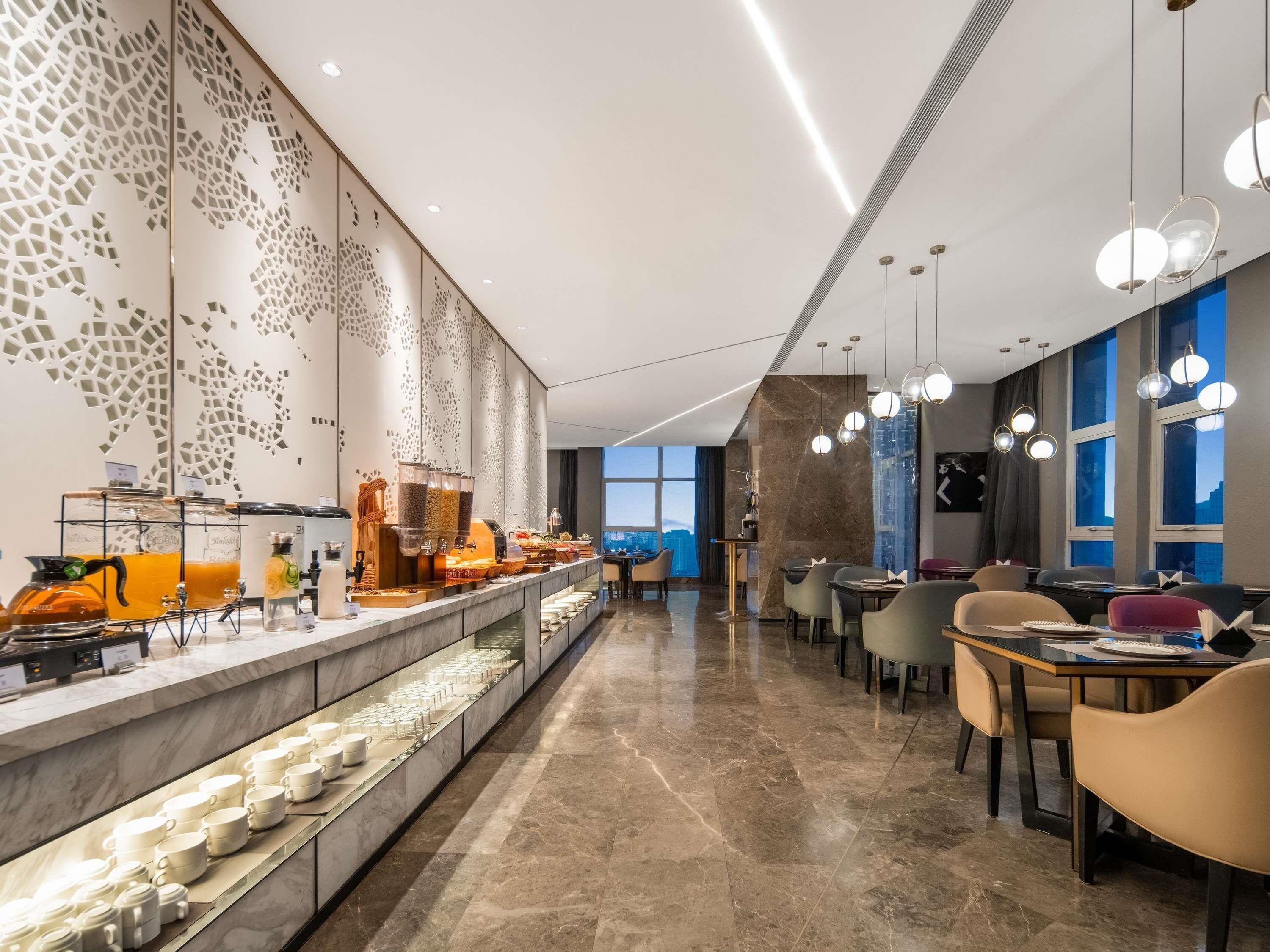 A modern hotel lobby with a long buffet counter, seating area, and large windows offering a city view.