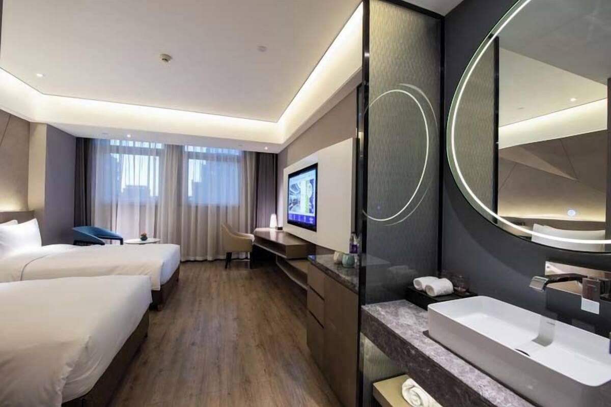 A hotel room with two beds, a desk with a TV, a large mirror, and a bathroom with a sink.