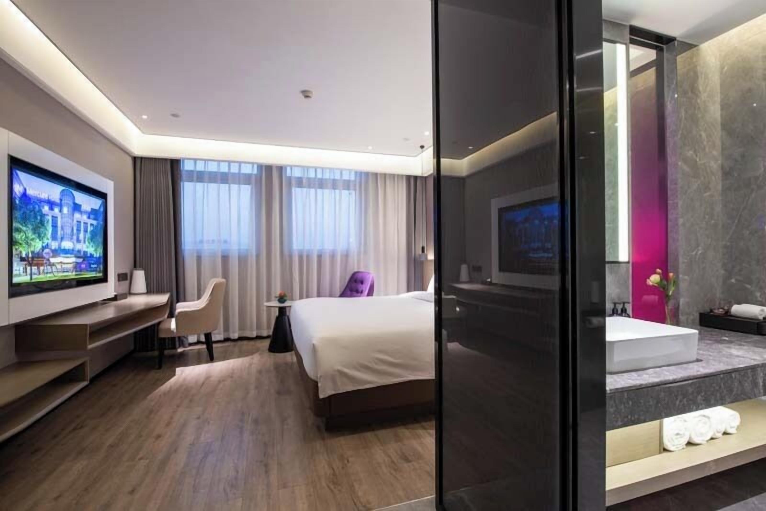 Una habitación de hotel moderna con una televisión de pantalla plana, una cama, una zona de estar con una silla, una mesita pequeña y un ventanal grande con cortinas.