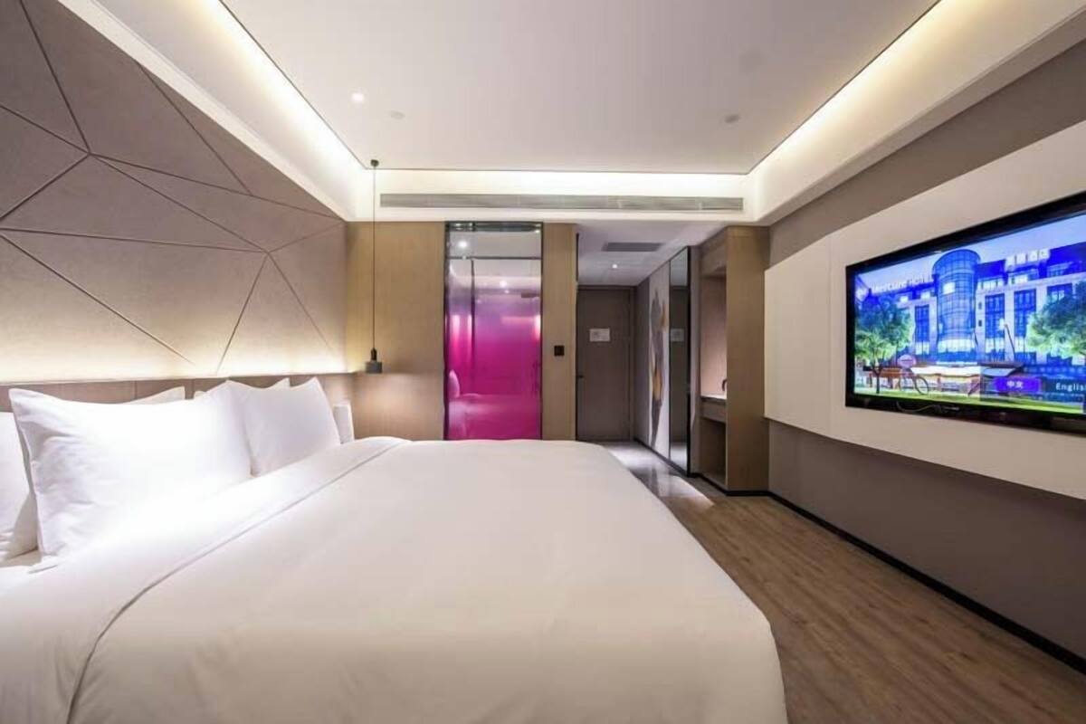 A modern hotel room with a large bed, a flat-screen TV mounted on the wall, and a view of the hotel's exterior through a glass door.