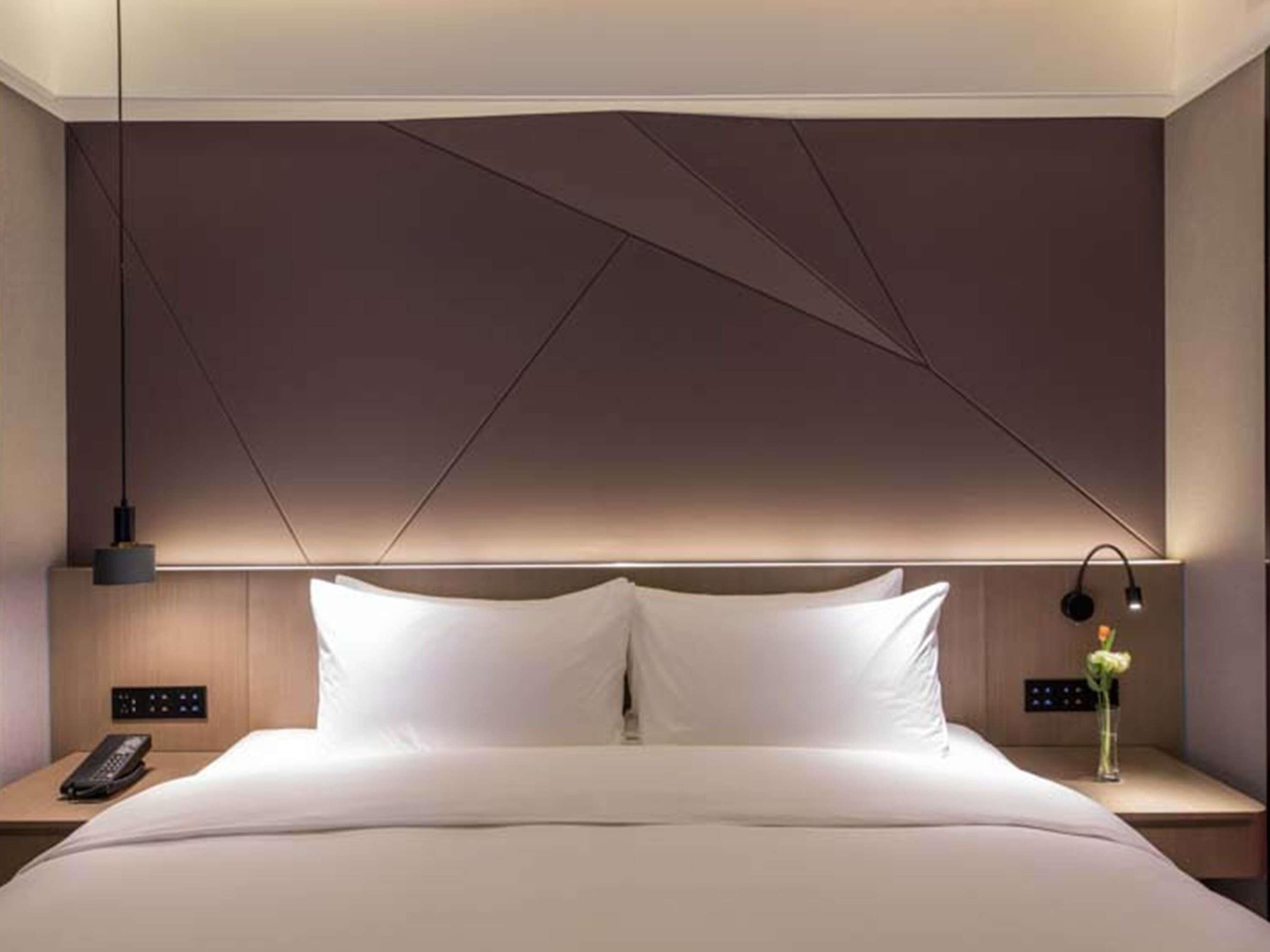 A modern hotel room with a large bed, bedside tables, a nightstand, a lamp, and a wall-mounted control panel.