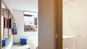 A modern hotel room with a wooden wardrobe, a bed, a blue ottoman, and a window with a city view. - ibis Budget Bogota Marly (Bogotá)