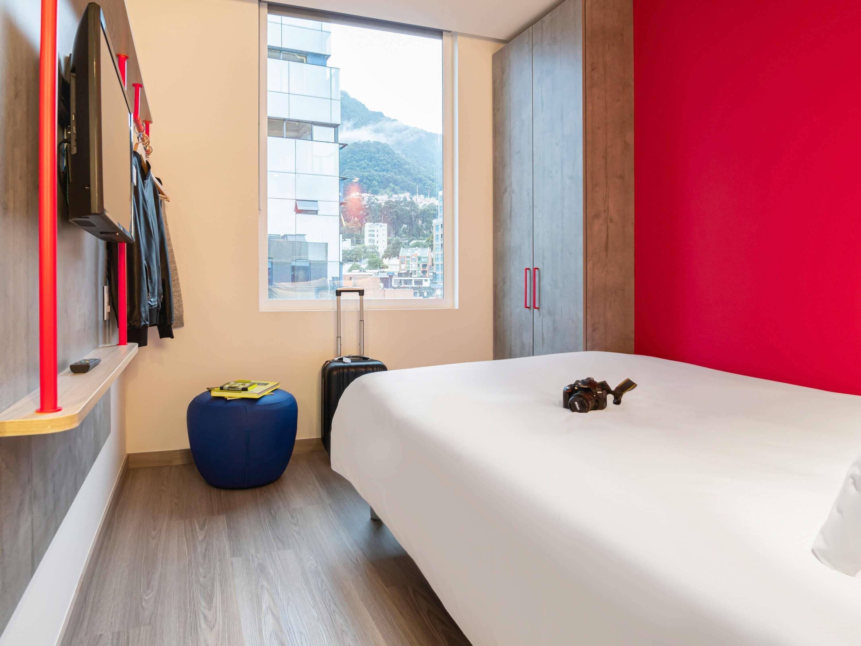 Een moderne hotelkamer met een bed, een koffer, een blauwe poef, een garderobe en een raam met uitzicht over de stad.