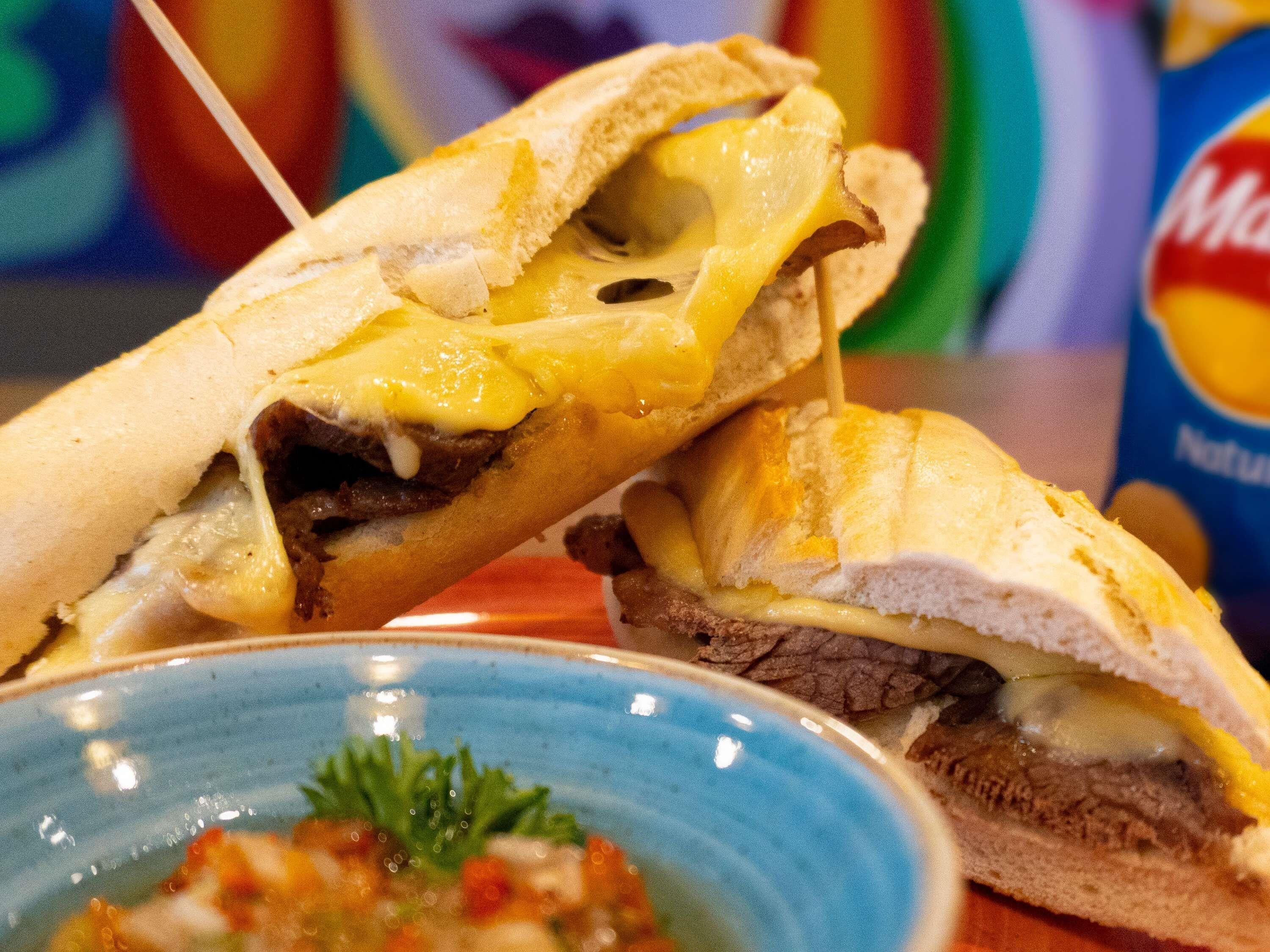 Un sandwich au fromage fondu et à la viande, une assiette bleue agrémentée d’une garniture, et un fond coloré.