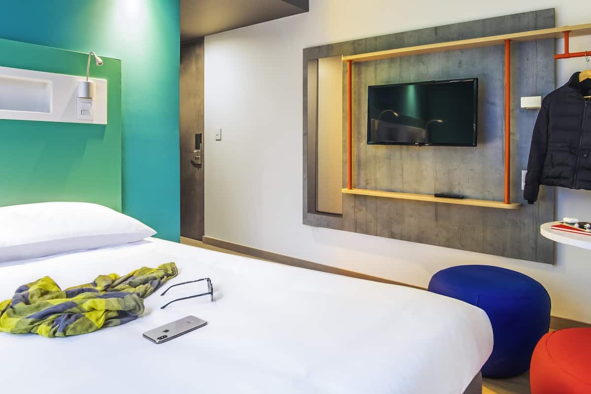 Ein modernes Hotelzimmer mit einem Bett, einem an einer grauen Wand montierten Fernseher, einem kleinen Tisch mit einer Fernbedienung und einem blau-roten Stuhl.