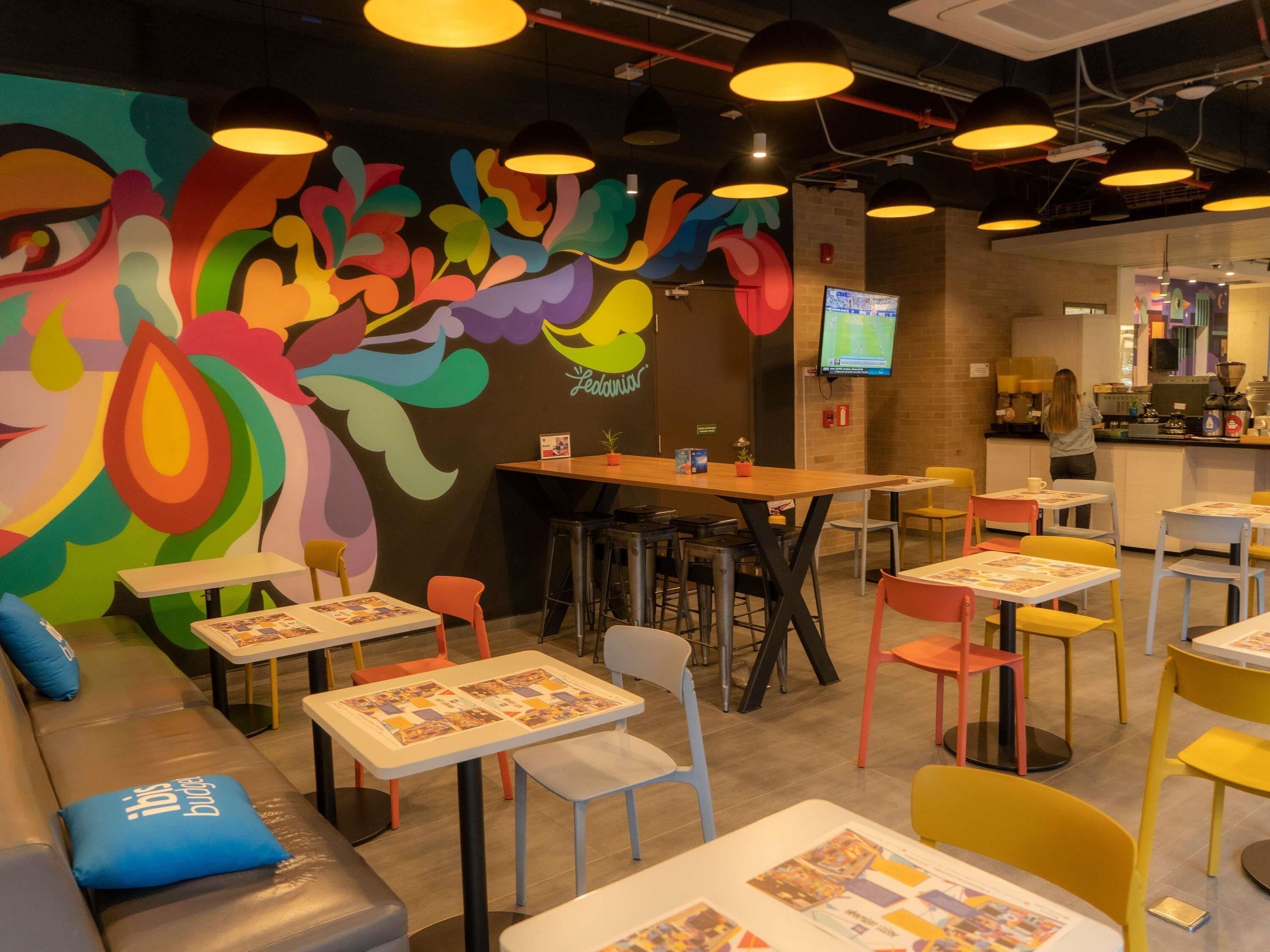 Un café animé, décoré de peintures murales colorées, offrant différents types de sièges, notamment des tables et des chaises, et dont la cuisine est visible.