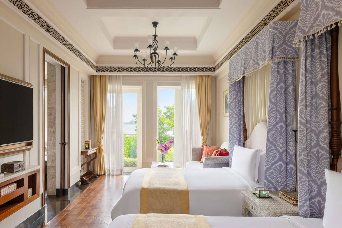 A hotel room with two beds, a television, a chandelier, and a view of greenery.
