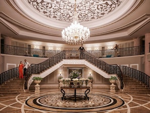 A grand lobby with a central staircase, ornate ceiling, and chandeliers.