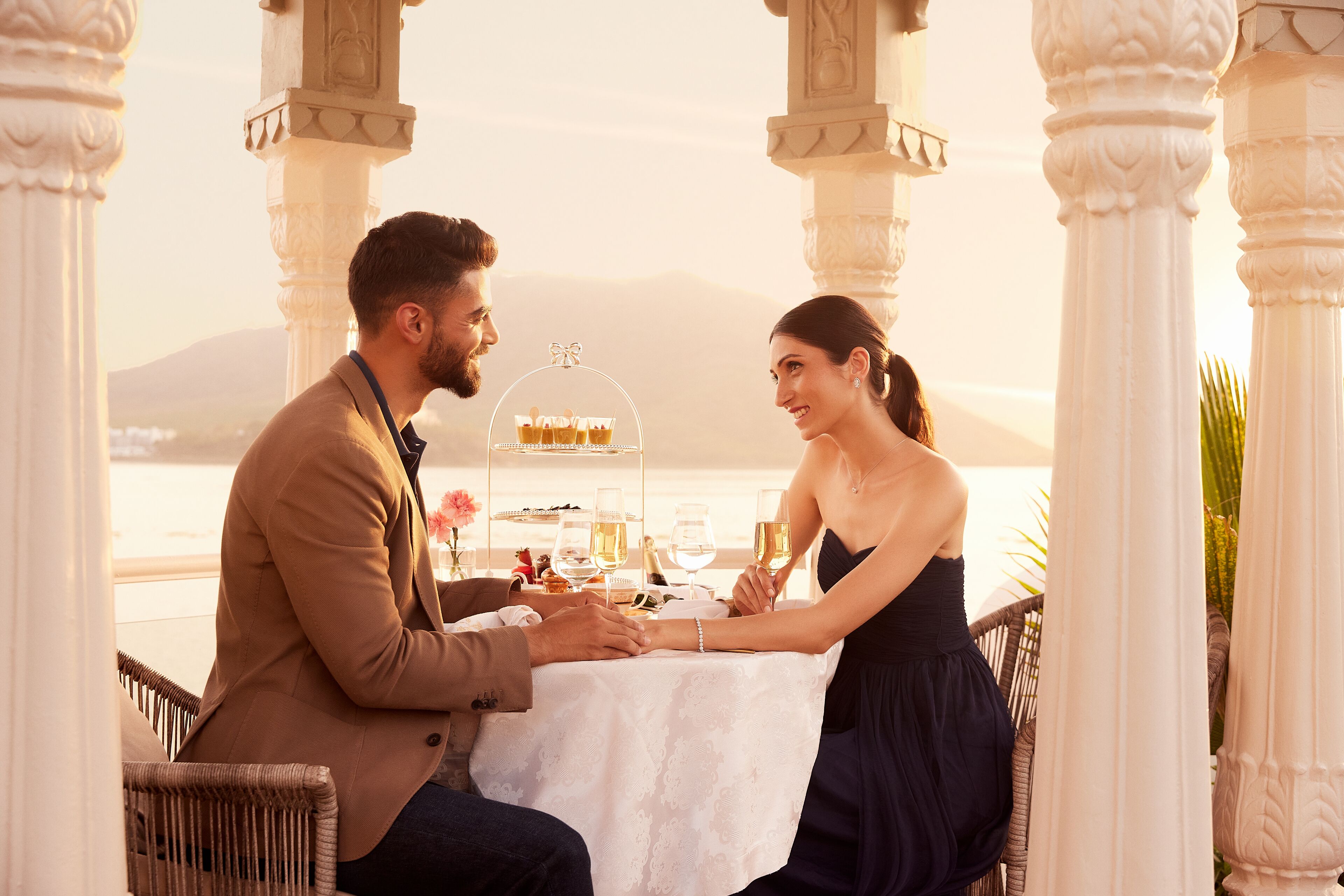 A couple dining at a table with a dessert display, surrounded by ornate columns and a view of a distant landscape.