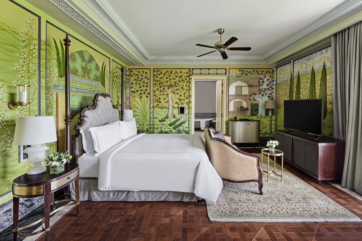 A hotel room with a large bed, a television, a desk with a lamp, a chair, and a bathroom visible through an open door.