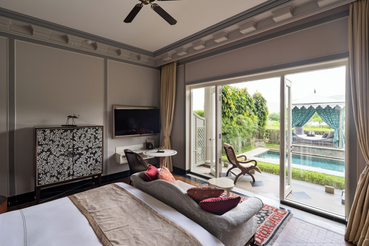 A modern hotel room with a large bed, a television, a decorative cabinet, a small table, and a view of a swimming pool and garden.