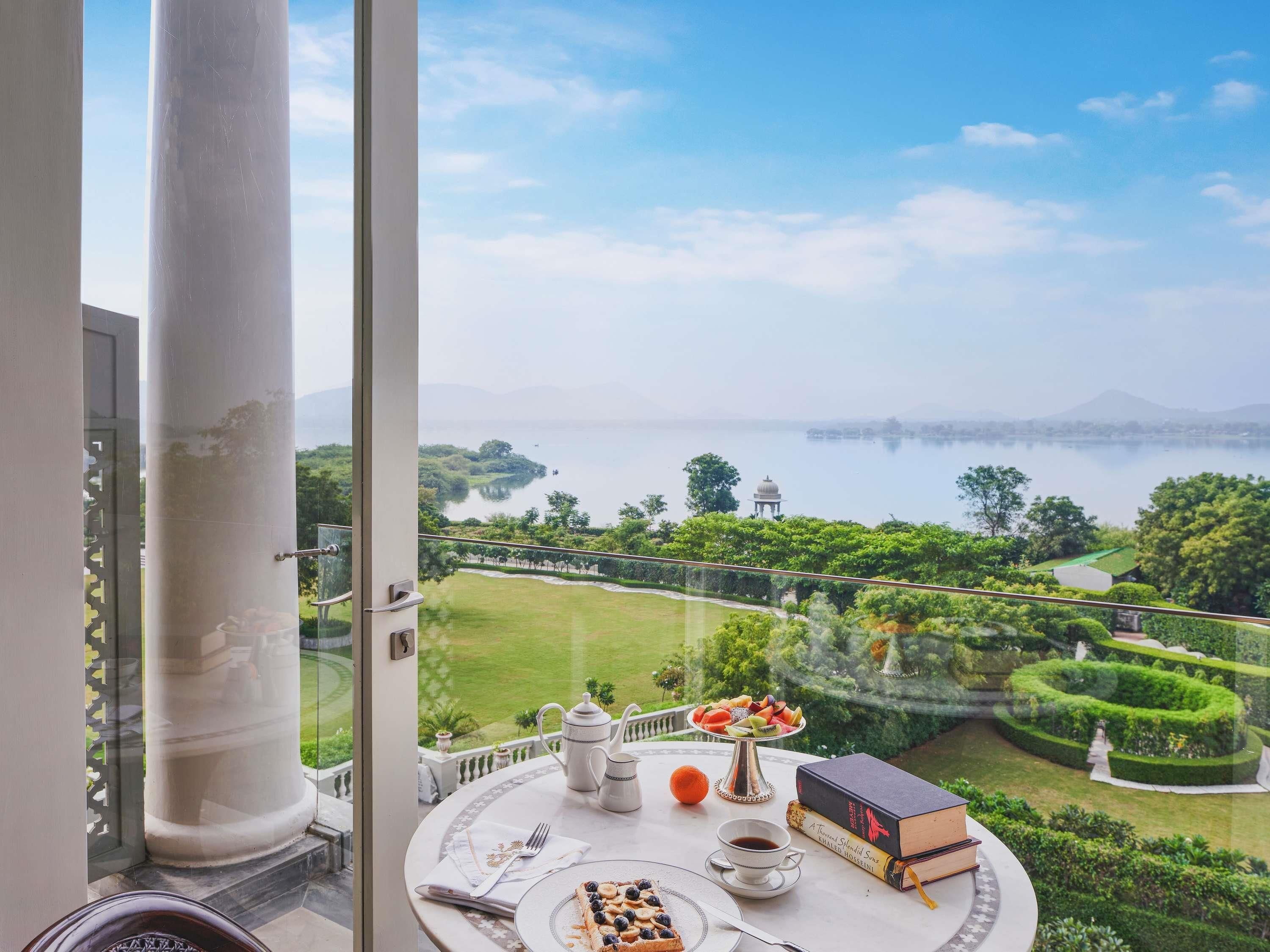 A balcony with a table set for tea, a book, and a view of a lake and garden.