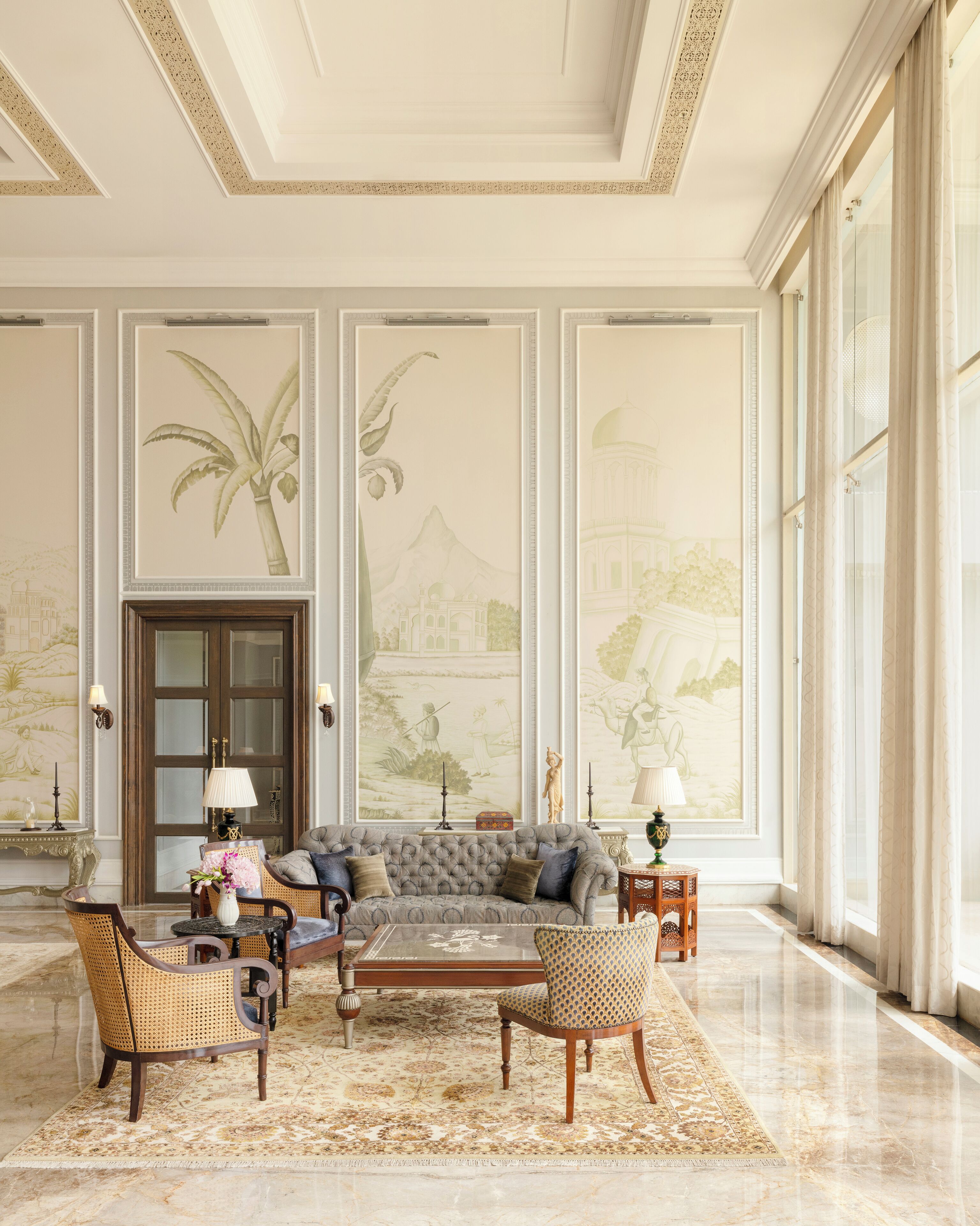 A spacious living room with a grand sofa, two armchairs, a coffee table, and a patterned rug. The room features ornate wall murals and large windows with curtains.