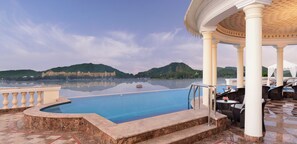 An infinity pool with a view of a hilltop building, a gazebo with columns, and outdoor seating.