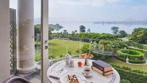 A balcony with a table set for tea, a book, and a view of a lake and garden. - Raffles Udaipur (Udaipur)