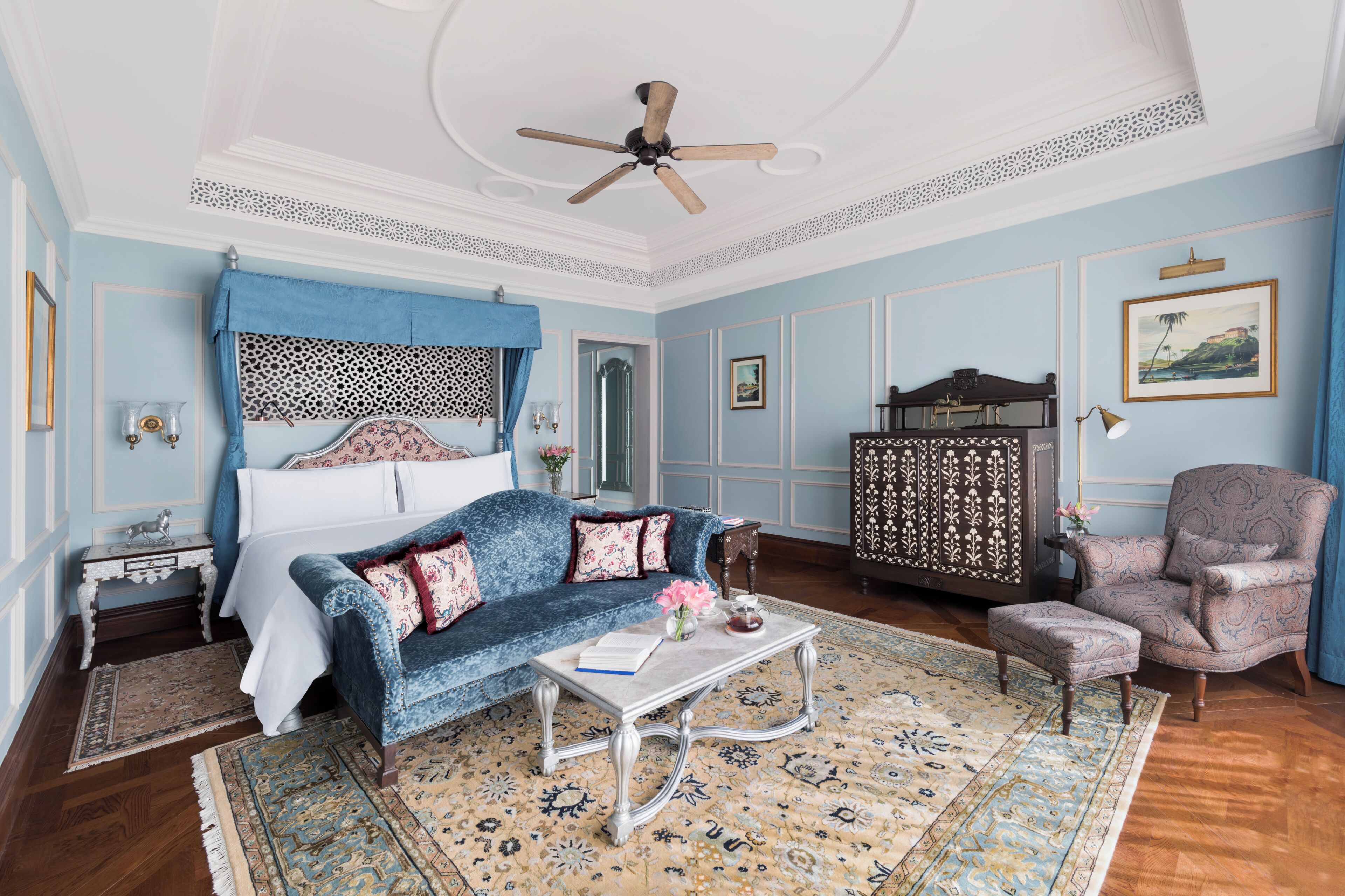 A room with a bed, a blue sofa, a patterned armchair, a coffee table, and a ceiling fan.