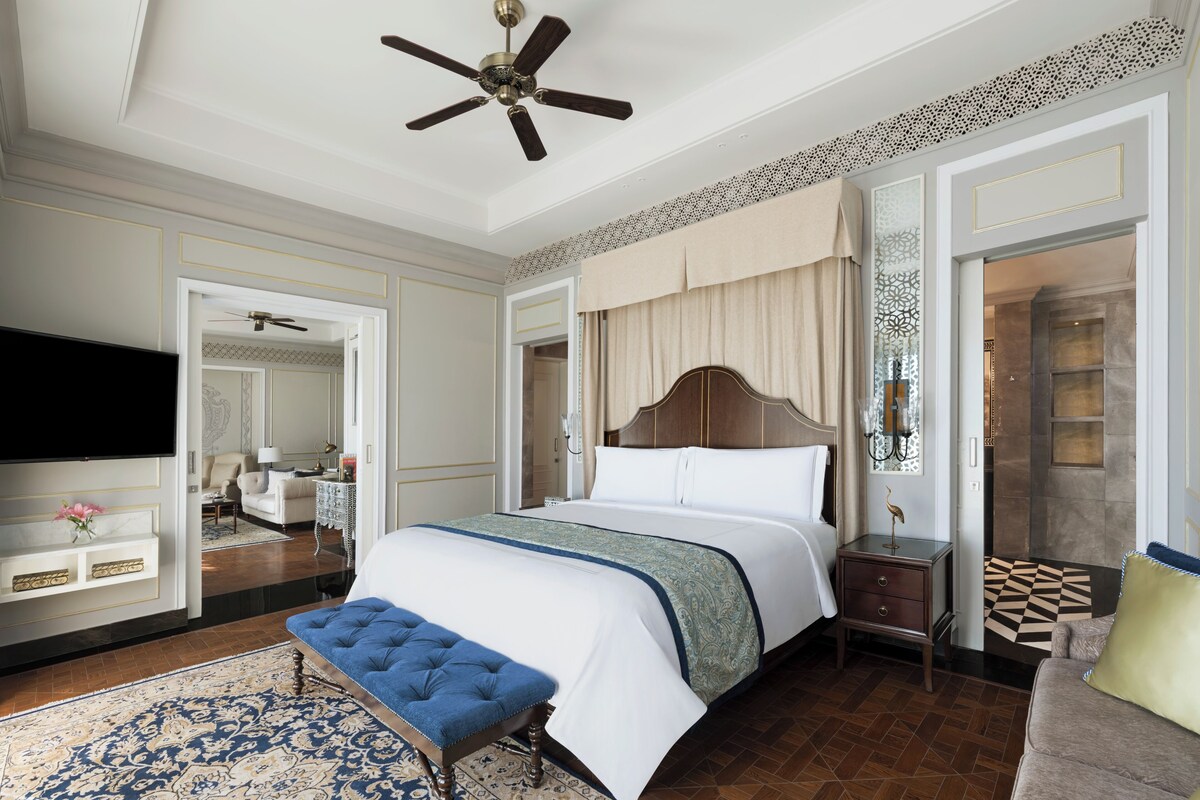 A hotel room with a large bed, a ceiling fan, a TV, a bedside table, a bench, and a patterned rug.
