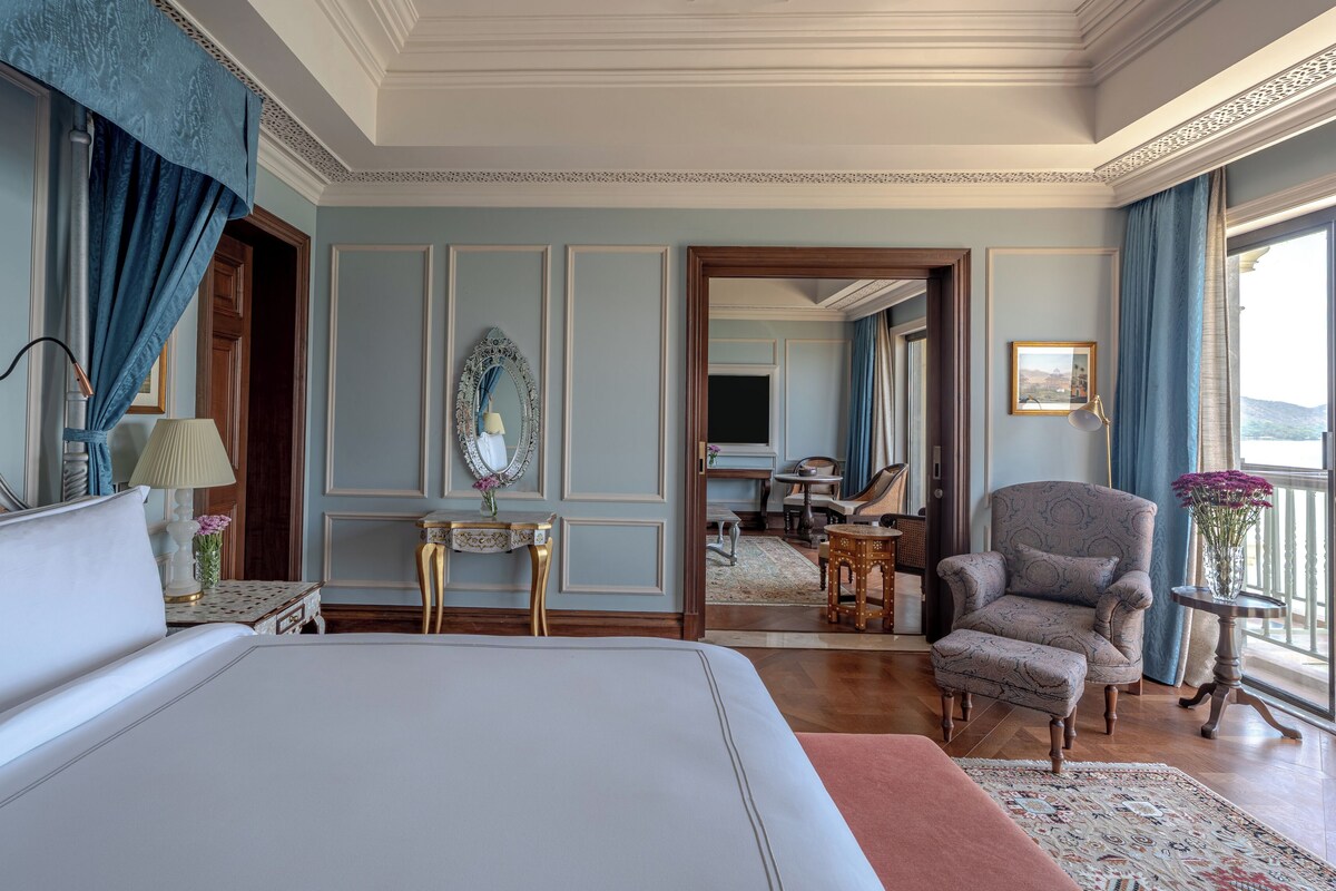 A spacious bedroom with a large bed, a sitting area with a chair and table, and a view of a balcony.