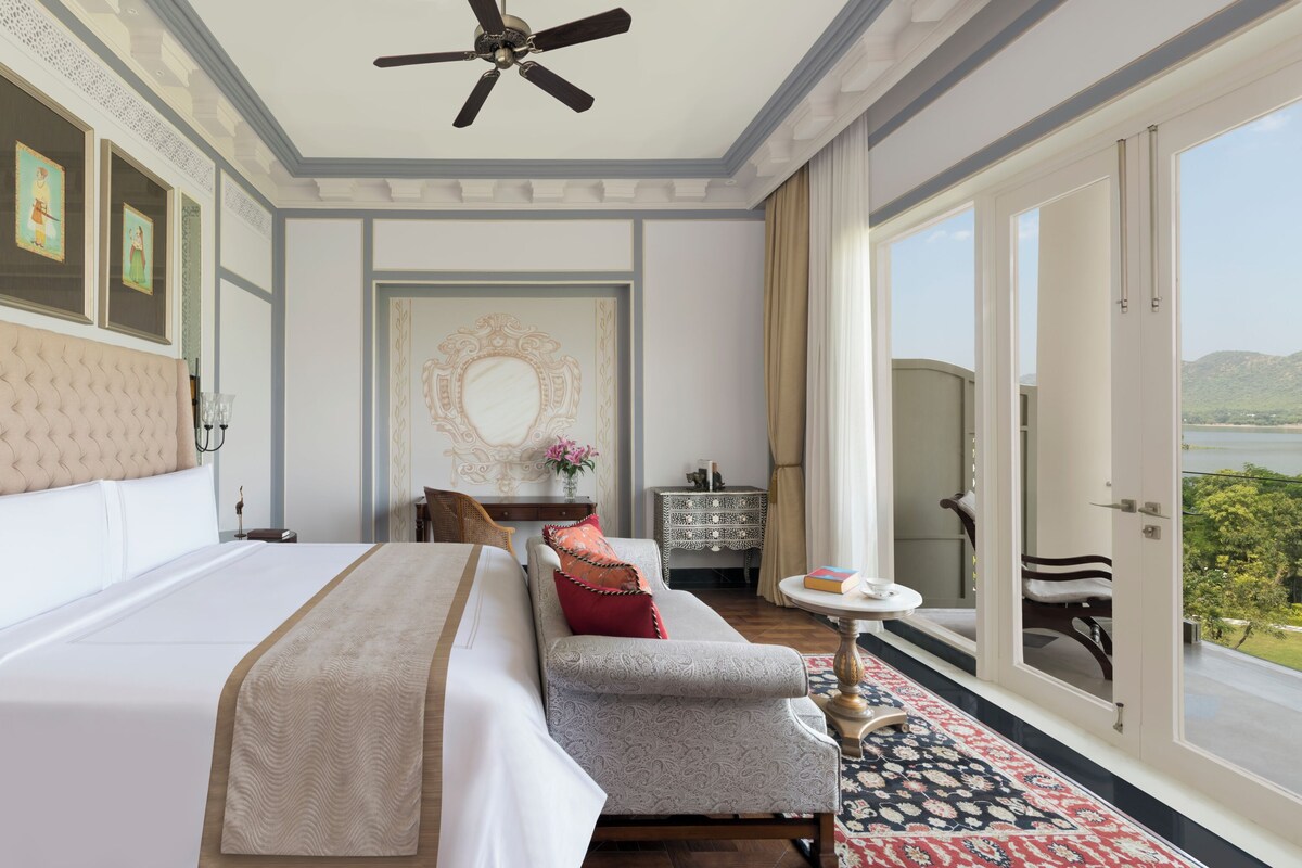 A hotel room with a large bed, a sofa, a small table, a ceiling fan, and a view of the outdoors.