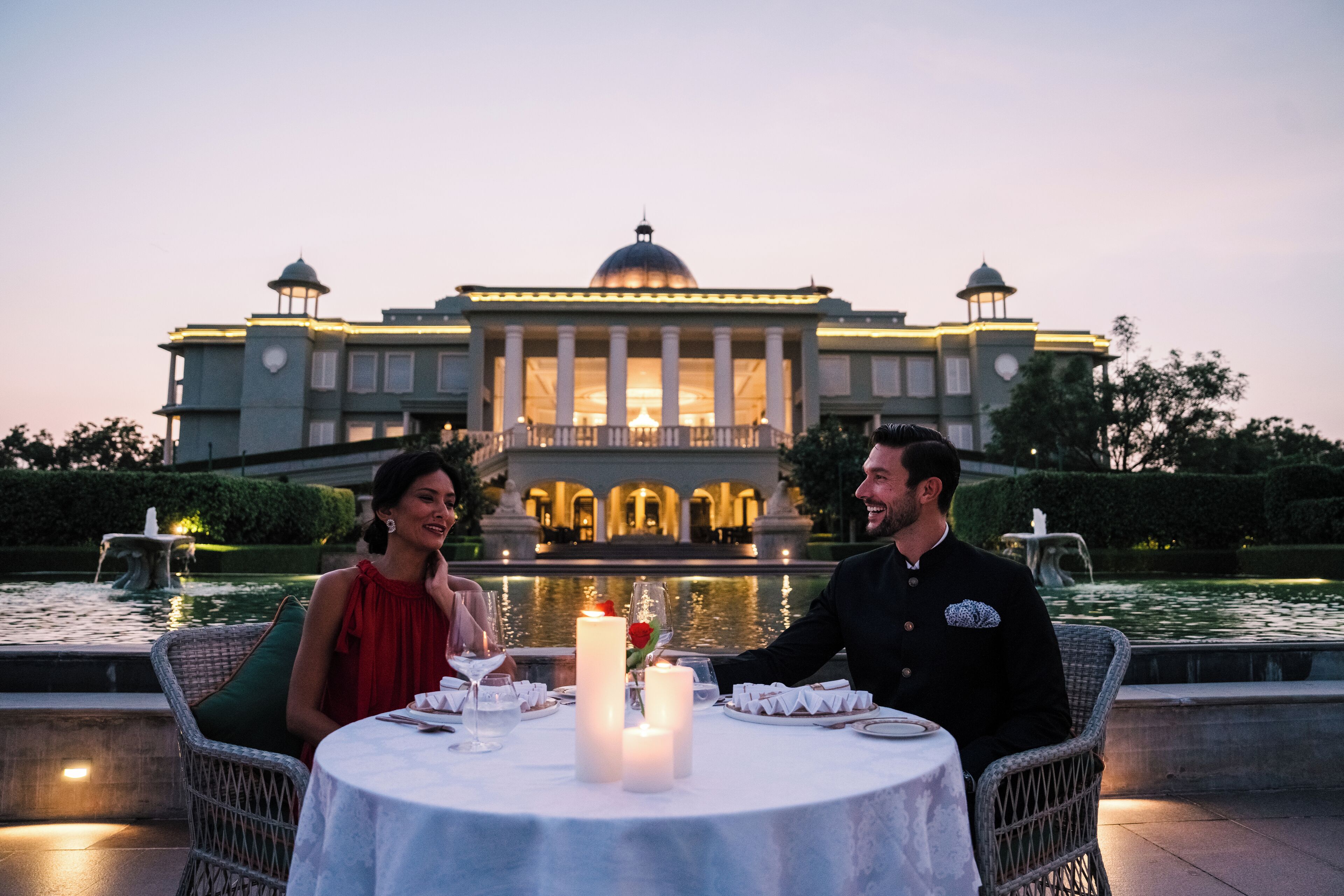 Un couple est assis à une table en plein air, éclairée par des bougies, devant un magnifique bâtiment orné d’une fontaine.