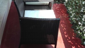 Et sæt af en fletstol og et bord på en terrasse med røde fliser, metalgelænder og grøn vegetation.