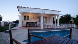 A poolside area with a covered patio, a wooden table, and a white fence.