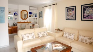 A living room with a beige sofa set, a glass coffee table, and a wooden side table. There is a dining area with a table and chairs in the background.