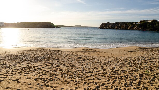 A beach with footprints in the sand, a clear sky, and buildings in the background.
