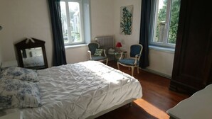 Un dormitorio con cama, dos sillas, una mesita, un radiador y una ventana con cortinas.