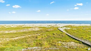 Un paisaje costero con una playa arenosa, dunas cubiertas de hierba y un paseo marítimo de madera que lleva a la playa.