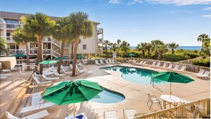 A pool area with lounge chairs and umbrellas, a building with balconies, and a view of the ocean.