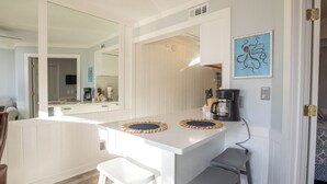 A compact kitchen with a white countertop, two stools, a coffee maker, and a large mirror.