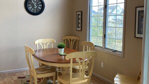 A dining area with a wooden table and chairs, a clock on the wall, and a window with a view of houses. - Close to everything! Short walk to beach, boardwalk and restaurants (Wildwood)