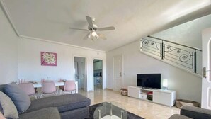 Sala de estar com sofá, televisão e ventoinha no teto. Escada com grades decorativas.