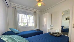 Quarto de hotel com duas camas de solteiro, ventoinha no teto, janela com cortinas, guarda-roupa, mesa de cabeceira com abajur e espelho na parede.