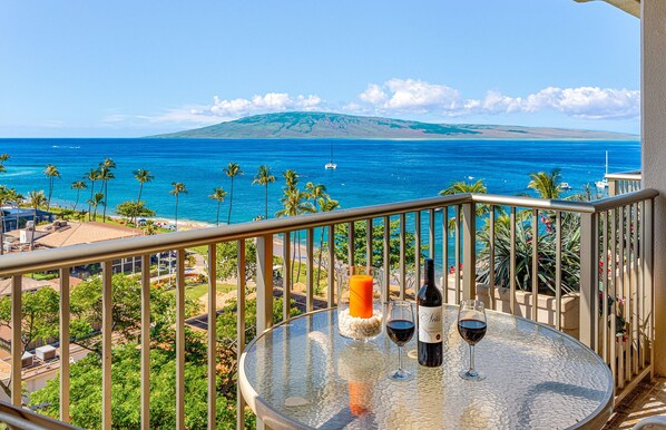 A balcony with a glass table, two wine glasses, and a candle, overlooking a coastal view with palm trees and a distant mountain.