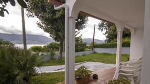 Um alpendre coberto com vista para um corpo d'água e montanhas, rodeado por vegetação e plantas em vasos.