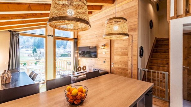 A modern kitchen with wooden cabinetry, a large island with a fruit bowl, pendant lights, and a view of the outdoors. - SAPELLI - Design apartment in brand-new residency (Chamonix-Mont-Blanc)