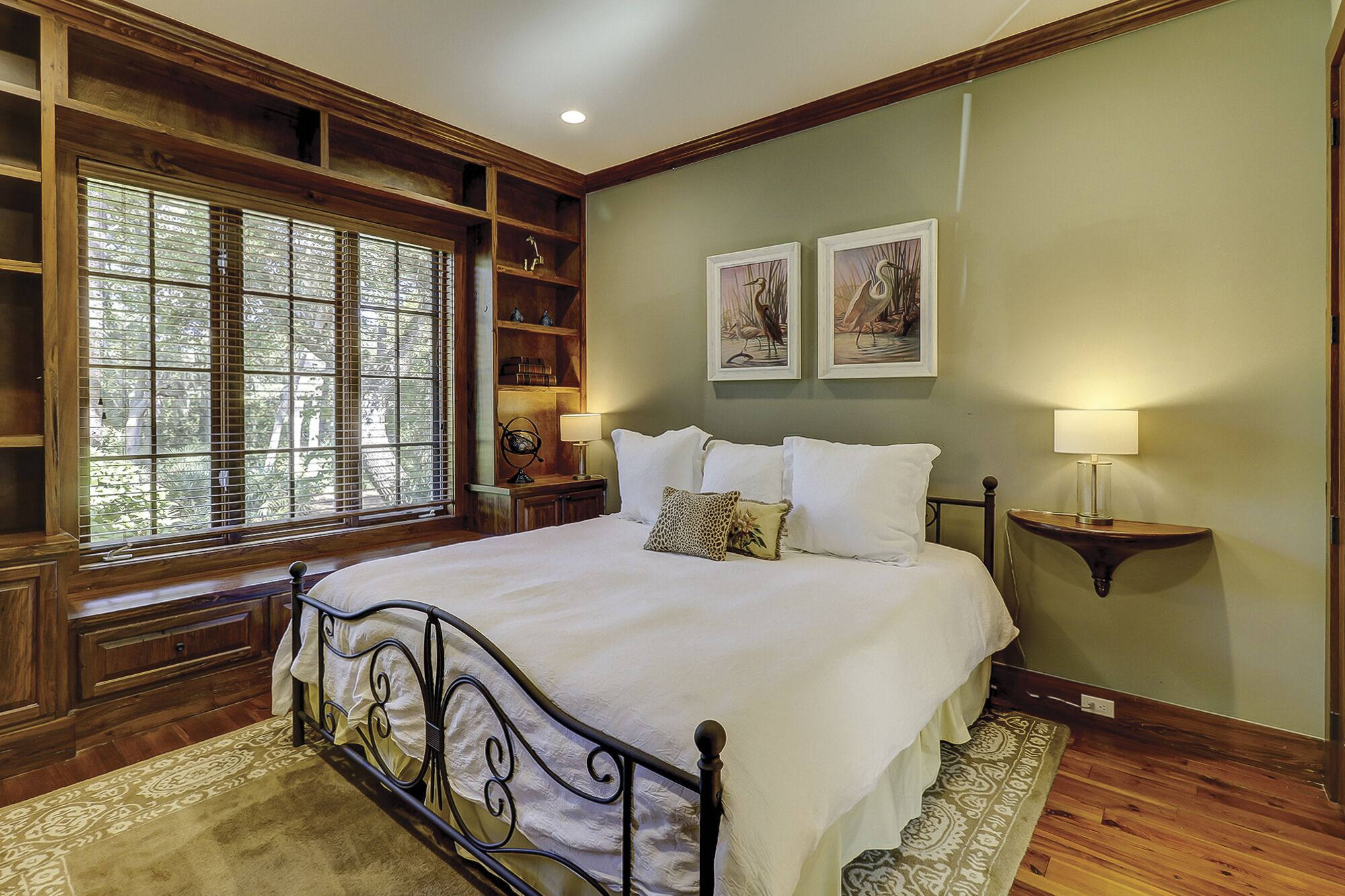 A bedroom with a large bed, wooden headboard, built-in shelving, and a window with blinds.