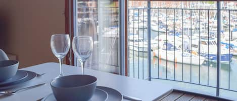 Mesa de comedor con platos, tazones y copas de vino dispuesta para dos personas, con vista a un puerto deportivo con barcos.