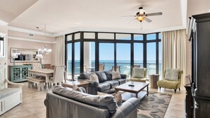 A spacious living area with a large window overlooking the ocean, a ceiling fan, and a dining area with a green cabinet. - The Oasis 409 | Spacious, Lazy River, Poolside Grill, New Splash Pad, Pool with Slide! (Orange Beach)