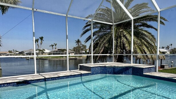 A clear swimming pool with a tiled edge, surrounded by a metal fence and palm trees. - Contemporary waterfront pool home with Gulf access (CAPE CORAL)