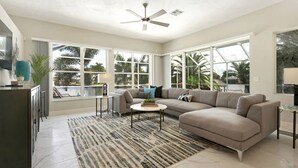 A spacious living room with a large sectional sofa, a coffee table, and a view of a pool and palm trees through large windows. - Contemporary waterfront pool home with Gulf access (CAPE CORAL)