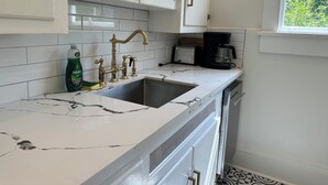 Una cocina con armarios blancos, encimera de mármol, lavamanos, cafetera y una ventana con vegetación al exterior.