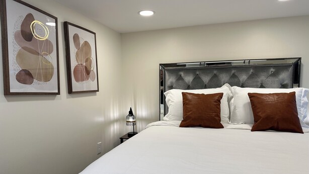Una cama bien hecha con almohadas blancas y marrones, un cabecero capitonado y dos cuadros abstractos en la pared.
