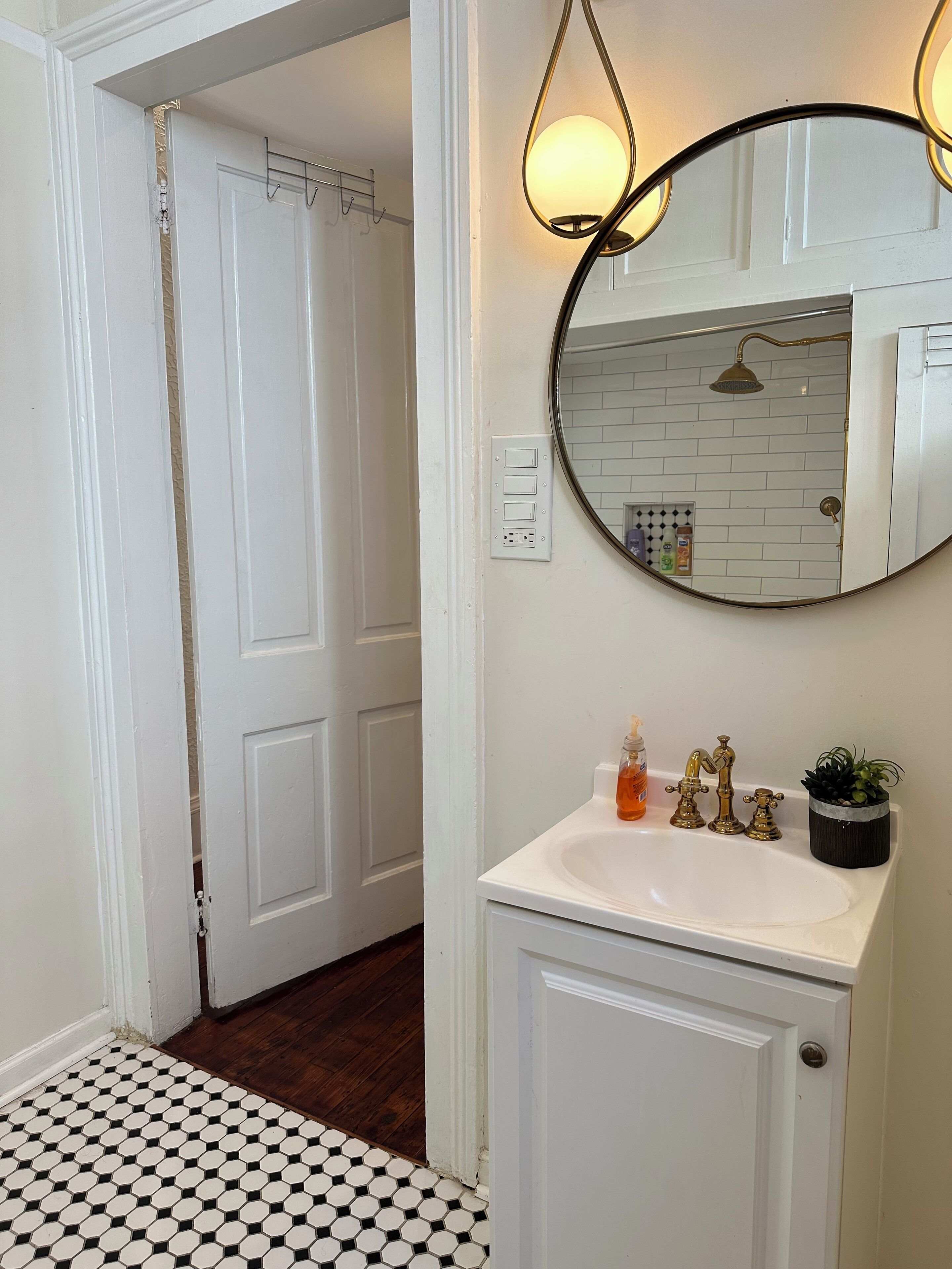 Une salle de bain avec un lavabo blanc, un miroir rond et une douche visible au fond.