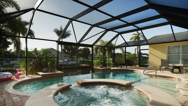 An indoor swimming pool with a hot tub, surrounded by a covered area with a metal frame and glass walls, and a patio area with chairs and a table. - Waterfront home with a Billiard table pool and spa (CAPE CORAL)