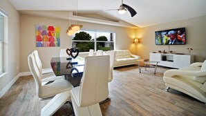 A modern living room with a dining area, featuring a glass table, white leather chairs, a sofa, a TV mounted on the wall, and a ceiling fan. - Waterfront home with a Billiard table pool and spa (CAPE CORAL)
