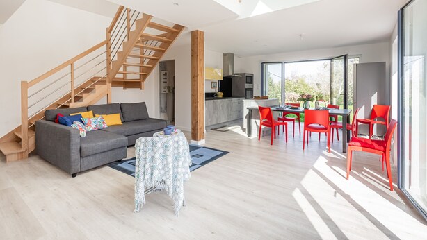 Un vaste espace de vie comprenant un canap é gris, un escalier en bois et un coin repas avec des chaises rouges.