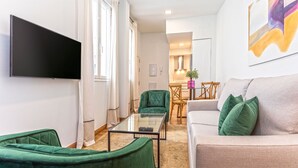 Un salon doté d’un téléviseur à écran plat, de fauteuils recouverts d’un tissu vert, d’une table basse en verre et d’un canapé agrémenté de coussins verts.